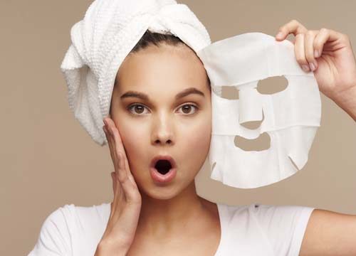 Woman applying a sheet face mask on a tan background